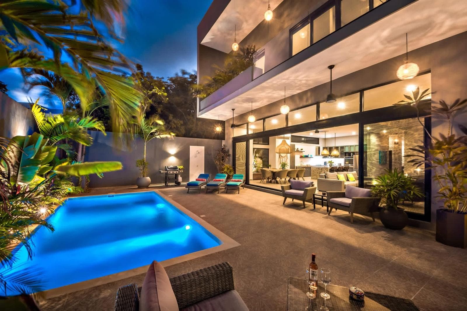 Villa at dusk with a lit pool, open living room and tropical garden